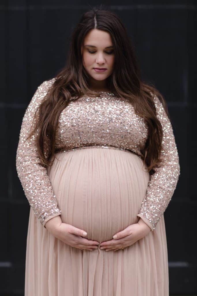 plus size pregnant woman wearing gold maternity dress, holding her pregnant belly and looking down. 