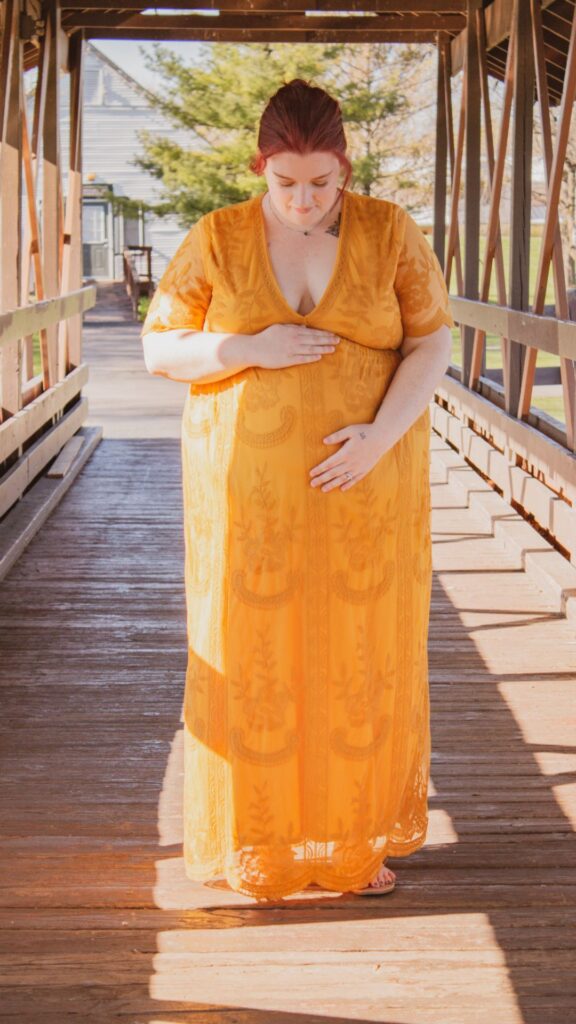 plus size pregnant woman standing on a bridge in a  yellow dress doing a forward-facing maternity pose